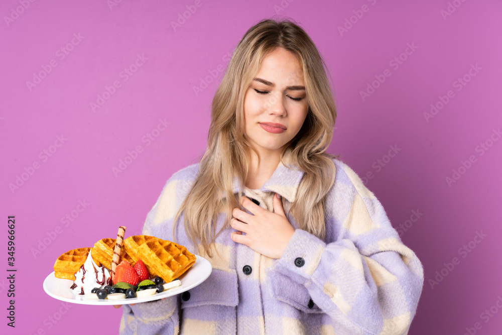 Teenager Russian girl holding waffles isolated on purple background having a pain in the heart