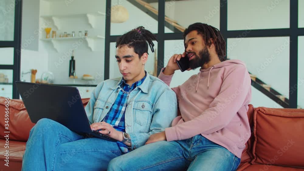 Cheerful stylish multiracial male friends with smart technology devices spending leisure together in domestic room. Positive young multiethnic men with dreads enjoying freetime and chatting indoors. Cheerful stylish multiracial male friends with smart technology devices spending leisure together in domestic room. Positive young multiethnic men with dreads enjoying freetime and chatting indoors.