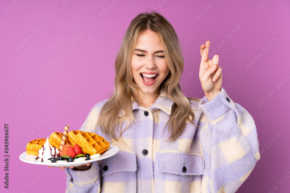 Teenager Russian girl holding waffles isolated on purple background with fingers crossing Teenager Russian girl holding waffles isolated on purple background with fingers crossing