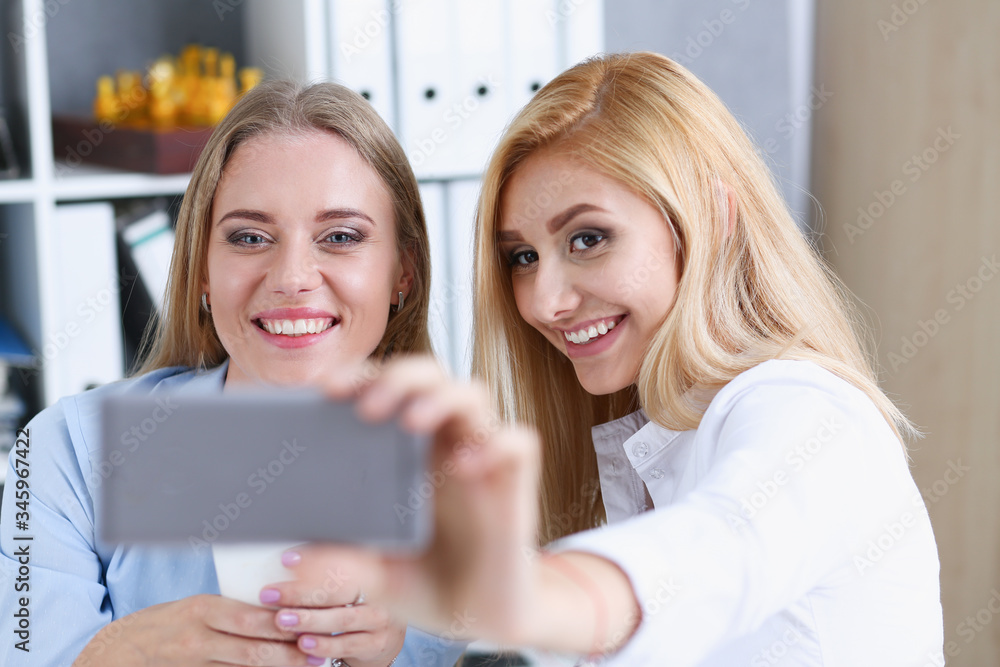 Two businesswomen in the office smile and do selfie on a smartphone during a break to have coffee Two businesswomen in the office smile and do selfie on a smartphone during a break to have coffee