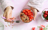 Concetto di colazione. Le mani della donna tiene una ciotola con fragole, cereali e yogurt su sfondo bianco con sfumature di rosa. Vista dall'alto