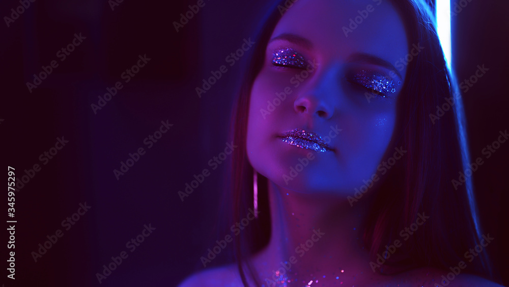 Neon light portrait. Creative fashion makeup. Woman with glitter lips closed eyes in blue purple glow. Neon light portrait. Creative fashion makeup. Woman with glitter lips closed eyes in blue purple glow.