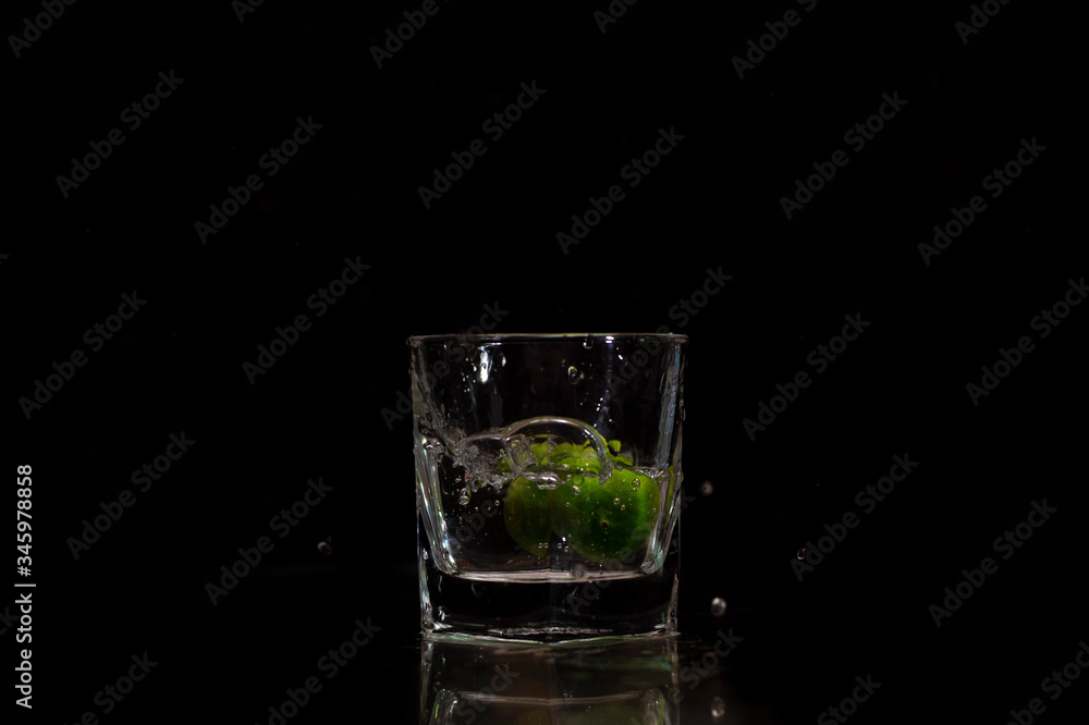Water splashes out of the garnished glass as a green fresh lemon slice immerses into the water. Water splashes out of the garnished glass as a green fresh lemon slice immerses into the water.