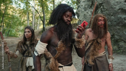 African wild tribe of aborigines walking in the woods. In focus black caveman talking on phone using miracle technology in prehistoric stone-age period. Fun and evolution. © Fractal Pictures