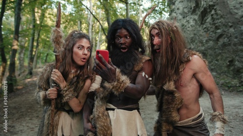 Valokuva Cheerful group of neanderthal hunters using modern technology smartphone cheering up walking outside cave in the jungle