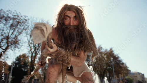 Εκτύπωση καμβά Aggressive wildman wearing animal skin using bow touching camera with fear or surprise discovering modern city