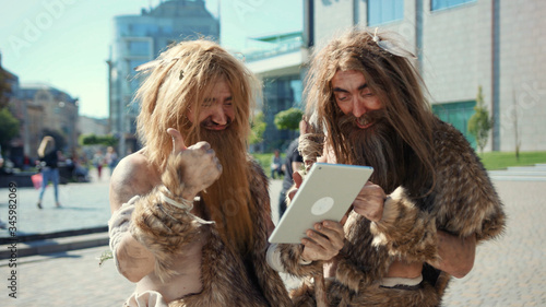 Konstfotografi Excited prehistoric bushmen of hunter-gatherers covered in fur browsing internet on tablet discovering technology in modern city