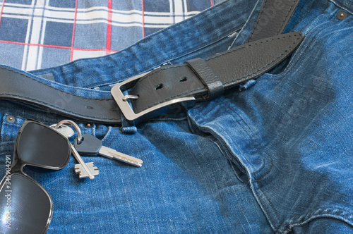 blue jeans with a black leather belt, a shiny buckle and two shiny keys next to dark sunglasses on a checkered background