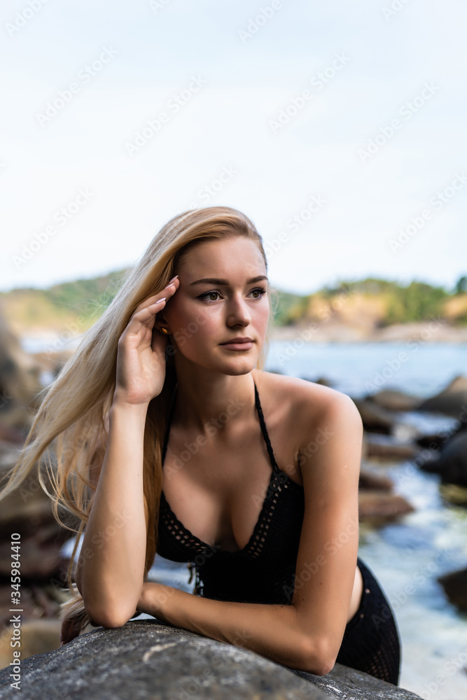 Young blonde woman in black bikini posing on a sand rocks near the sea Young blonde woman in black bikini posing on a sand rocks near the sea