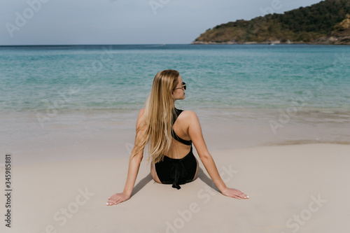 Rear view of young blonde woman wear in black bikini and sunglasses sitting on beach © F8  \ Suport Ukraine