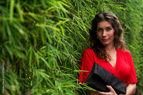 Fashionable beautiful brunette girl with python clutch on green natural background. Curly young woman in red dress holding leather snakeskin python bag. Stylish accessories.