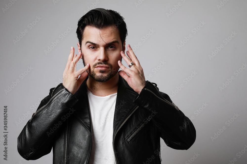 Portrait of brutal man with dark beard, looks aside, wears leather jacket, over blank studio background with free space. Portrait of brutal man with dark beard, looks aside, wears leather jacket, over blank studio background with free space.