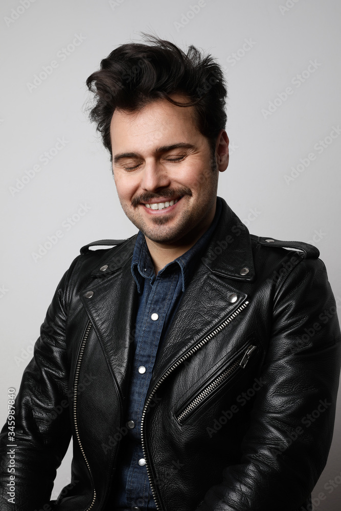 Funny bearded man wearing biker jacket, smiles strangely and looks at the camera with closed eyes. Vertical shot. Funny bearded man wearing biker jacket, smiles strangely and looks at the camera with closed eyes. Vertical shot.