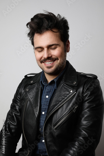 Funny bearded man wearing biker jacket, smiles strangely and looks at the camera with closed eyes. Vertical shot.