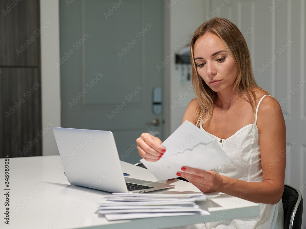 Fototapeta premium Worried unhappy female with laptop opening envelopes with bills