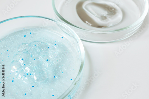 Close-up blue shower gel with scrub grain with bubbles and transparent shampoo smudge in glass petri dish with selective focus on white background. Concept laboratory tests and research