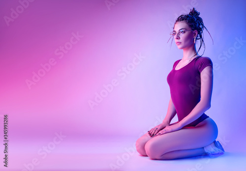 a girl dressed in a bodysuit with dreadlocks on her head is sitting on the floor on a white background in neon light