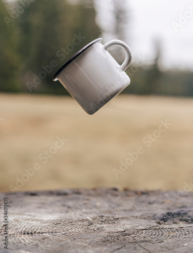 White enamel cup hanging in the air above a wooden ground. Metal camping cup flying in the air in the outdoors. Close up detail photo of a camping coffee mug. photo