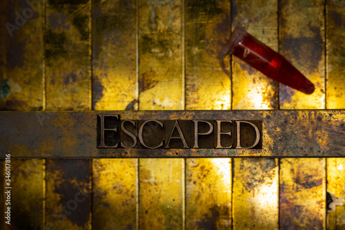 Photo of real authentic typeset letters forming Escaped text with red fluid filled laboratory vial on vintage textured grunge copper and gold background