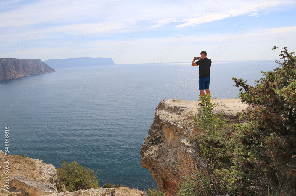 A man, a tourist stands on a high mountain and photographs the landscape, the sea on the phone. Beautiful sea coast, view from the cliff. Traveling alone. 