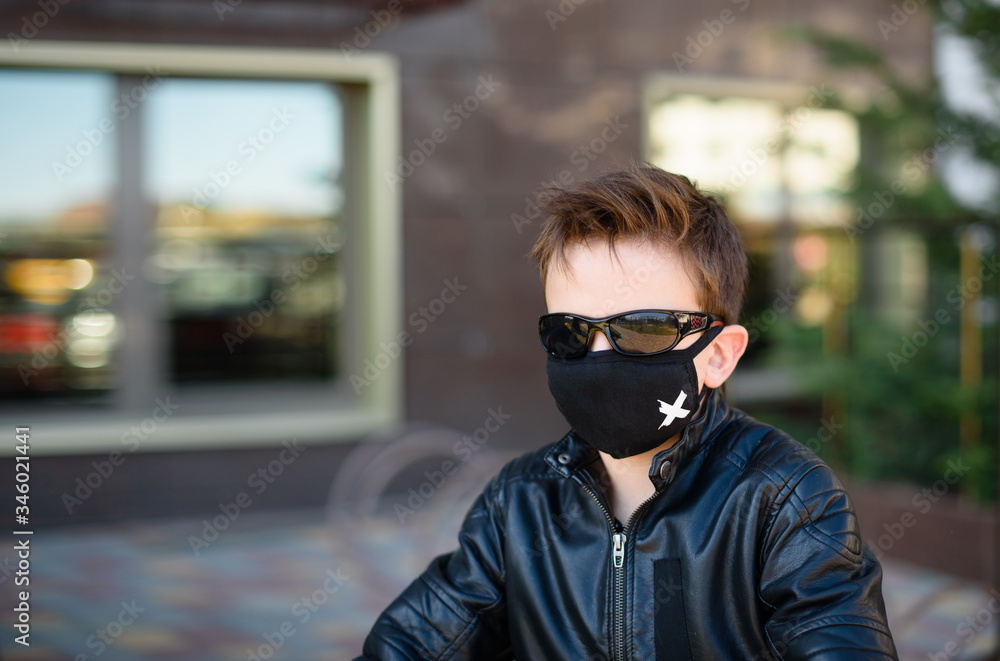 Boy in a leather jacket wearing a black hygyenic medical mask outdoors in the yard in black sunglasses, looking forward. White cross on a black mask Boy in a leather jacket wearing a black hygyenic medical mask outdoors in the yard in black sunglasses, looking forward. White cross on a black mask