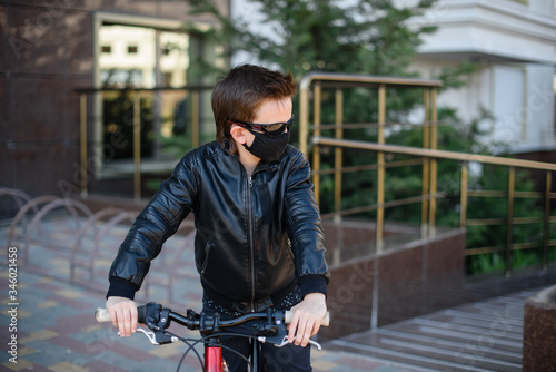 Boy in a leather jacket wearing a black hygyenic medical mask outdoors in the yard in black sunglasses, riding a bike. View from the side. Cancelled vacation, bad mood due to coronavirus covid-19