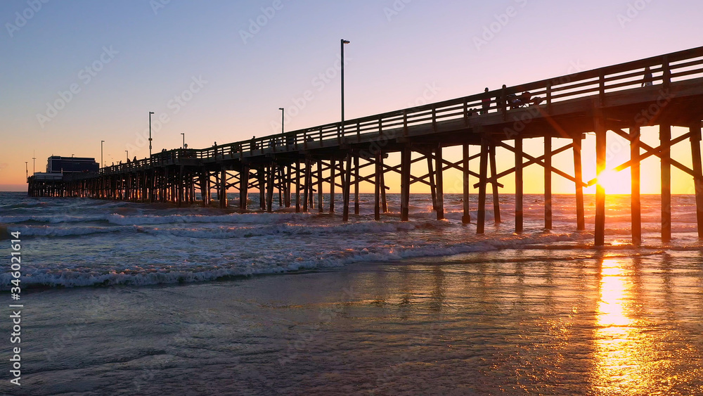 People walking on the Newport Beach pier during sunset as waves slowly roll in to the beach.