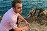 young handsome bearded guy wearing pink polo sitting on the hill on background of sea and horizon line, looking away