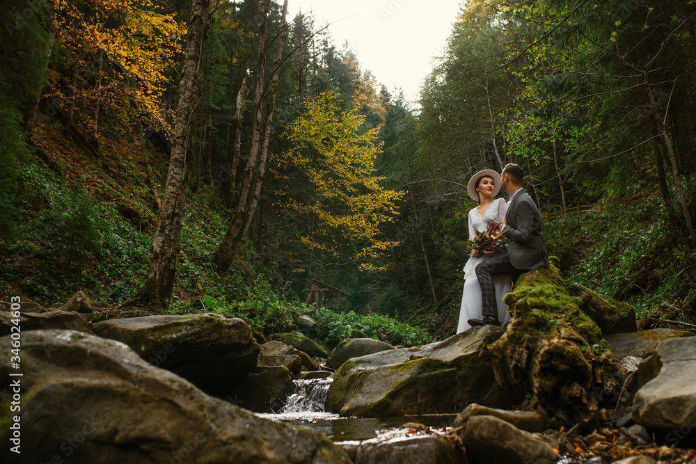 the bride and groom are sitting on a stone in the middle of a mountain river in the forest. Mountain wedding. Wedding in the forest. the bride and groom are sitting on a stone in the middle of a mountain river in the forest. Mountain wedding. Wedding in the forest.