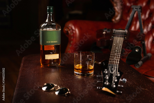 A bottle of Irish whiskey, with a glass, glasses, and a guitar on the table. Place for text or logo.