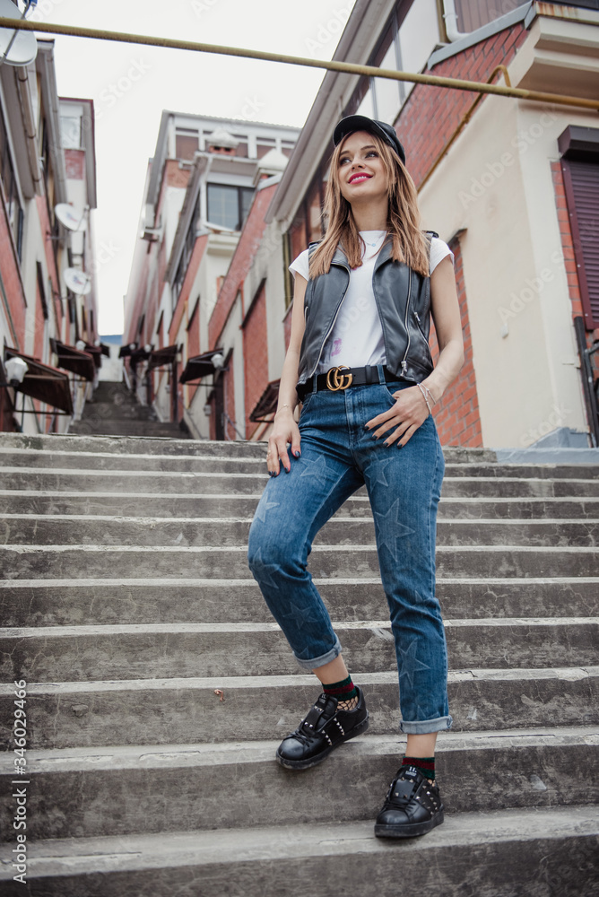 Beautiful in a leather vest, jeans with stars and a cap stands in the city on the stairs Beautiful in a leather vest, jeans with stars and a cap stands in the city on the stairs