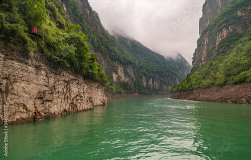 Wuchan, China - May 7, 2010: Dawu or Misty Gorge on Daning River. Bend in canyon with brown rocky cliffs, green foliage on top, emerald green water, and misty cloudscape. © Klodien