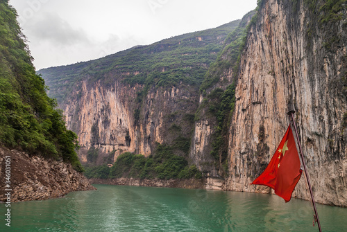 Wuchan, China - May 7, 2010: Dawu or Misty Gorge on Daning River. Chinese red flag in canyon with brown rocky cliffs, green foliage on top, emerald green water, and misty cloudscape. photo