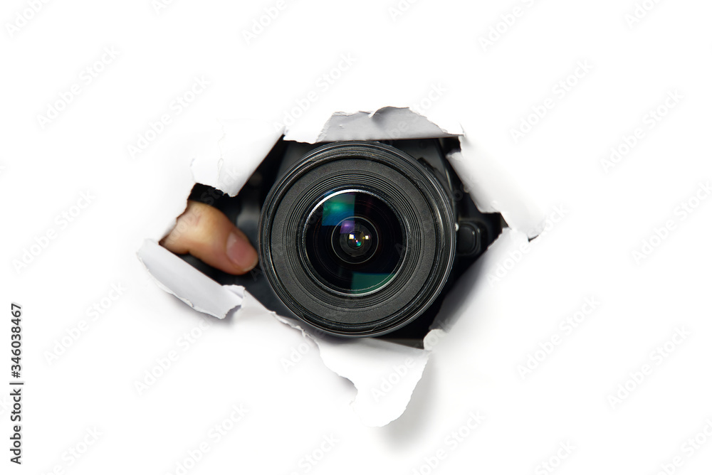 black camera with a telephoto lens that looks out through a hole in white paper. Concept of paparazzi, espionage, yellow press. camera lens looking through hole in wall.