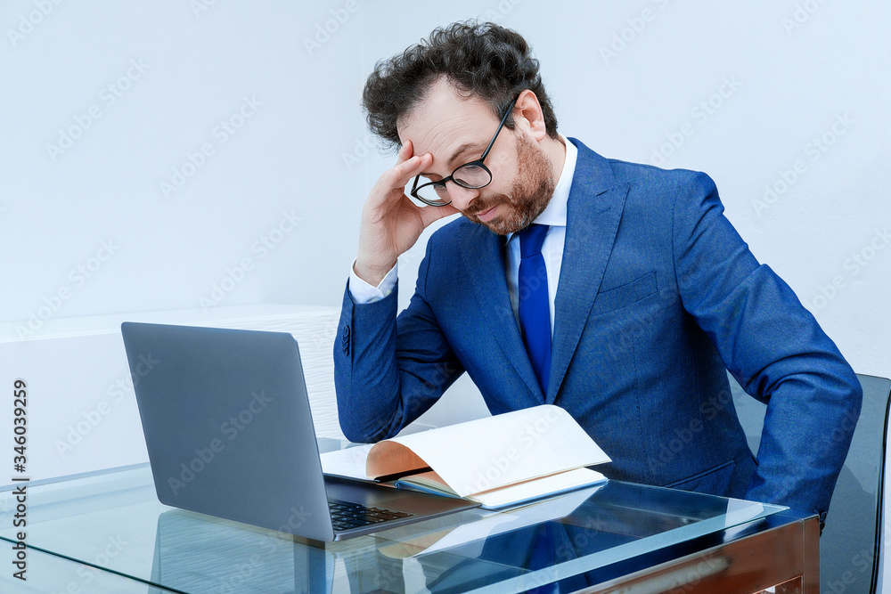 Businessman with thoughtful face in blue suit using laptop on office interior backdrop. Concept of come up with a new strategy, anticrisis plan Businessman with thoughtful face in blue suit using laptop on office interior backdrop. Concept of come up with a new strategy, anticrisis plan