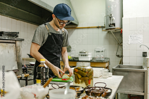 Fototapeta Naklejka Na Ścianę i Meble -  Man with glasses and a cap cooking in a restaurant kitchen with pots of food, oil and pans