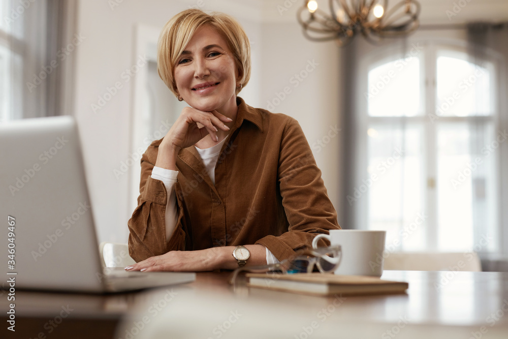 Female With Laptop. Senior Blonde Woman In Brown Jacket Sits At The Desk With Laptop And Cup. Businesswoman Homework Concept. Female With Laptop. Senior Blonde Woman In Brown Jacket Sits At The Desk With Laptop And Cup. Businesswoman Homework Concept.