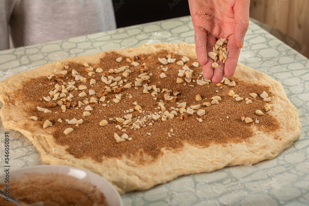 woman hand sprinkles peeled roasted peanuts on rolled dough with cinnamon. the action takes place in the kitchen, the dough on the table. near the dough saucer with cinnamon.