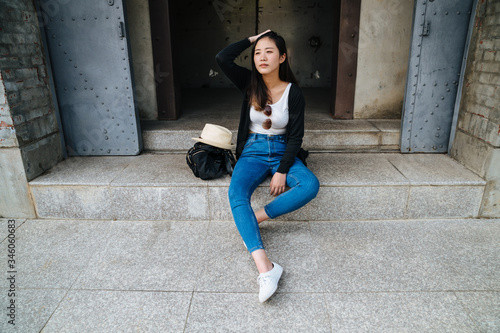 Young stylish woman in jeans and black light jacket resting on old factory entrance stairs. Attractive asian chinese girl flicks short hair sitting on steps and looking aside with backpack and hat