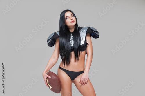 Beautiful sexy brunette female american football player in uniform posing with a ball isolated on grey background