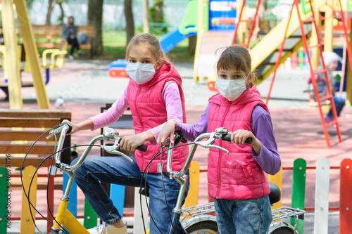 Two girls in medical masks ride bicycles in the playground