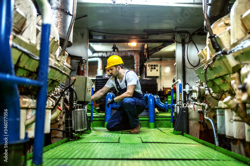 Worthy caucasian mechanic in overalls and with helmet kneeling inside ship and repairing engine.