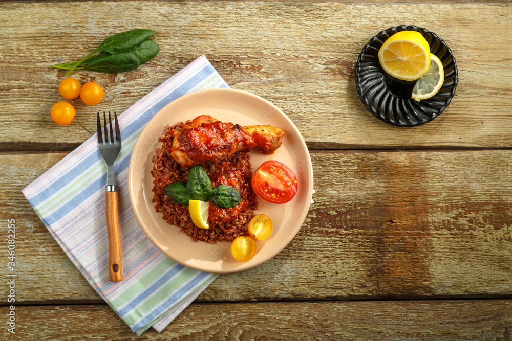 Chicken legs with red rice in tomato-garlic sauce on a plate on a wooden table on a linen napkin.
