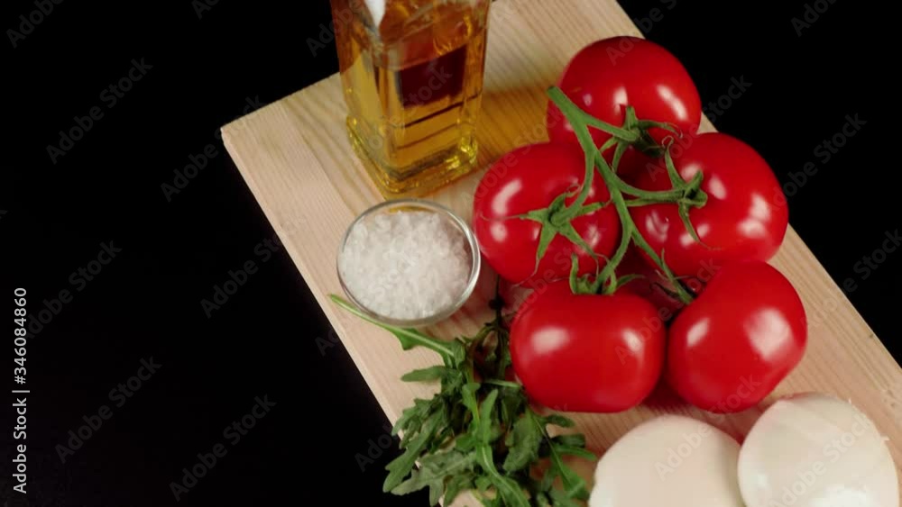 Modern caprese salad. Fresh juicy red tomatoes on a branch, mozzarella cheese, arugula leaves, olive oil and sea salt on a wooden board against black background. Camera slides down. Top view. Close up Modern caprese salad. Fresh juicy red tomatoes on a branch, mozzarella cheese, arugula leaves, olive oil and sea salt on a wooden board against black background. Camera slides down. Top view. Close up