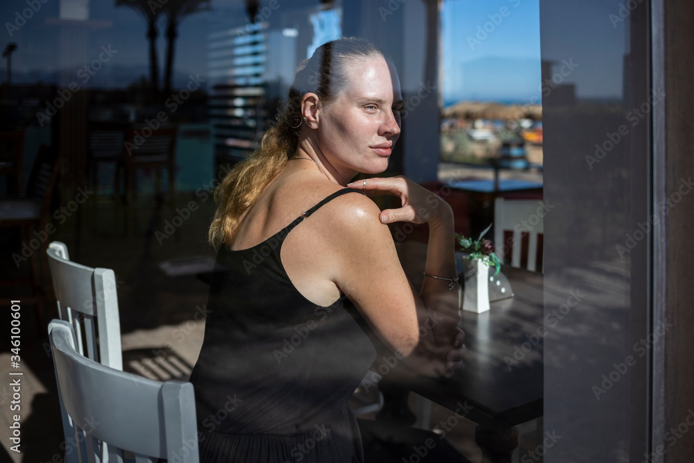 Portrait of a beautiful woman through a restaurant glass. A woman is sitting on a white chair in a black dress. In the background is a sea beach. The girl thought, dreams. The girl has silver jewelry.