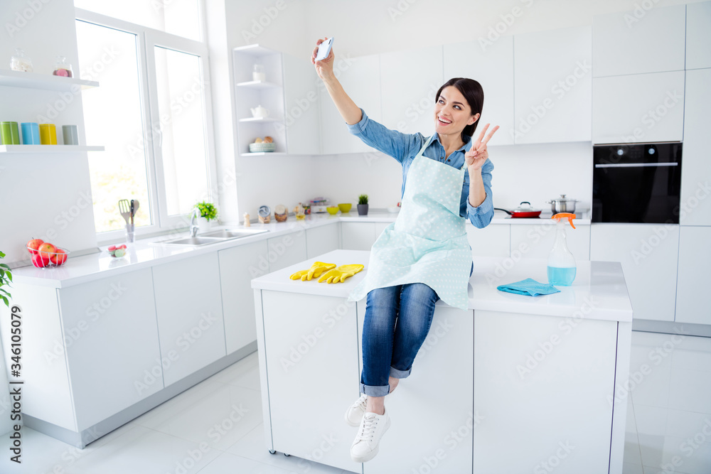 Full length photo of positive woman house wife corona virus protection prevention influencer sit table make selfie smartphone show v-sign in house indoors kitchen Full length photo of positive woman house wife corona virus protection prevention influencer sit table make selfie smartphone show v-sign in house indoors kitchen