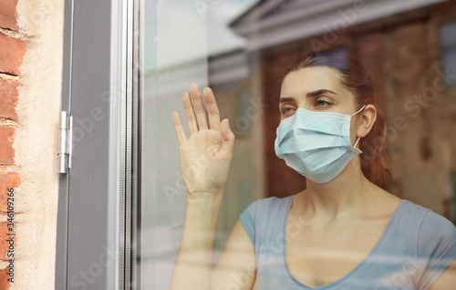 health, safety and pandemic concept - sick young woman wearing protective medical mask looking through window at home