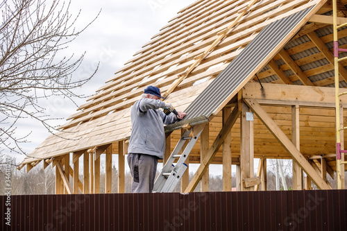 Senior gray-haired Construction man using a screwdriver, fastens a roofing sheet to wooden rafters on the roof of a country house under construction. Physical activity of the seniors.
