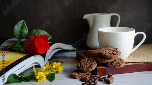 Coffee cups and chocolate chip baked cookies with red roses on books on a wooden table