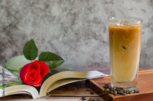 Ice coffee in a tall glass with cream poured over and coffee beans on a old rustic wooden table andbeautiful red rose flowers on the book.cold summer drink on a dark wooden background with copy space. photo